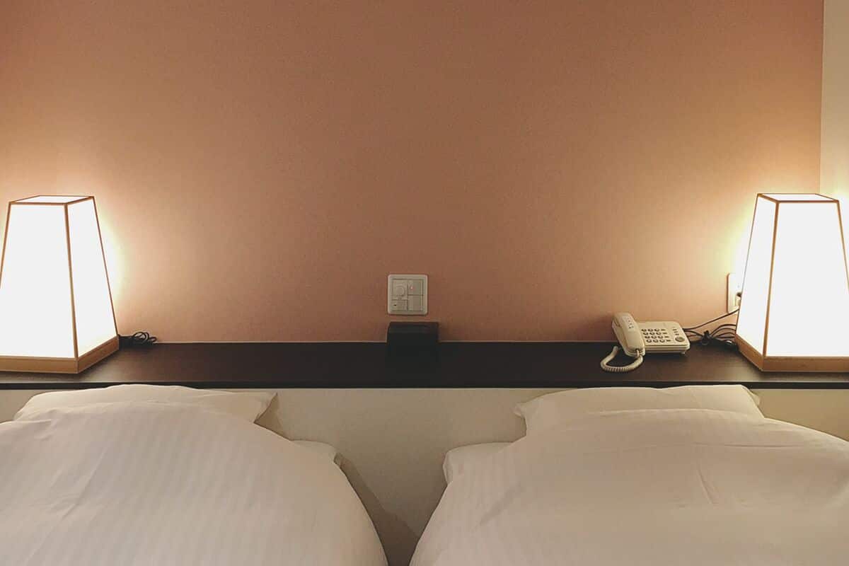 A hotel room with two beds, a bedside table, a lamp, a phone, and a thermostat.