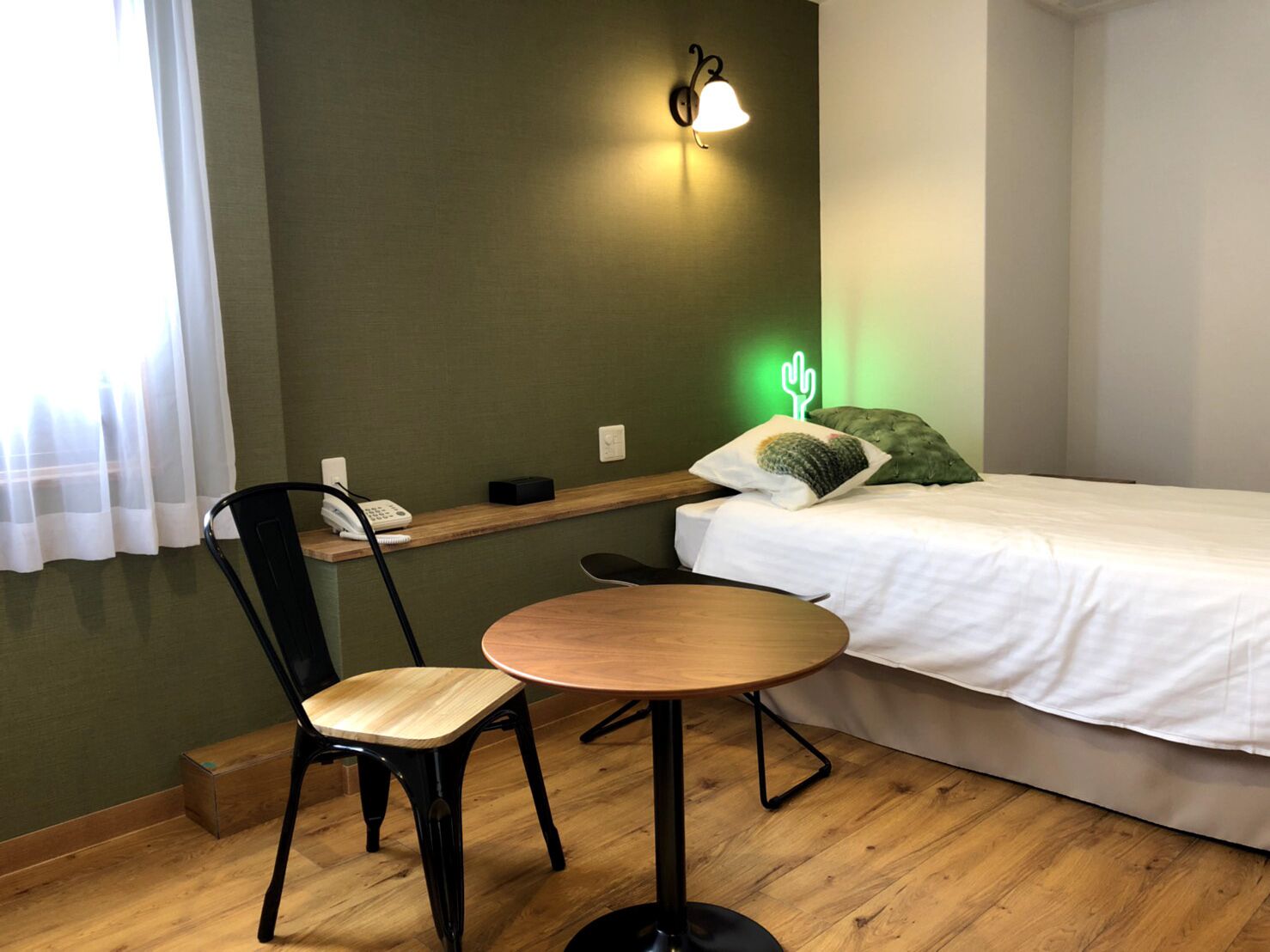 Una habitación pequeña y ordenada con una cama, una sola silla, una mesita y un teléfono en la pared, además de un cojín con forma de cactus.