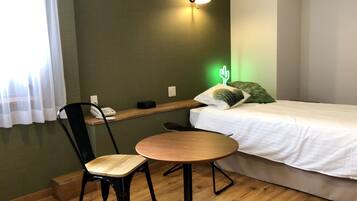 Una habitación pequeña y ordenada con una cama, una sola silla, una mesita y un teléfono en la pared, además de un cojín con forma de cactus.