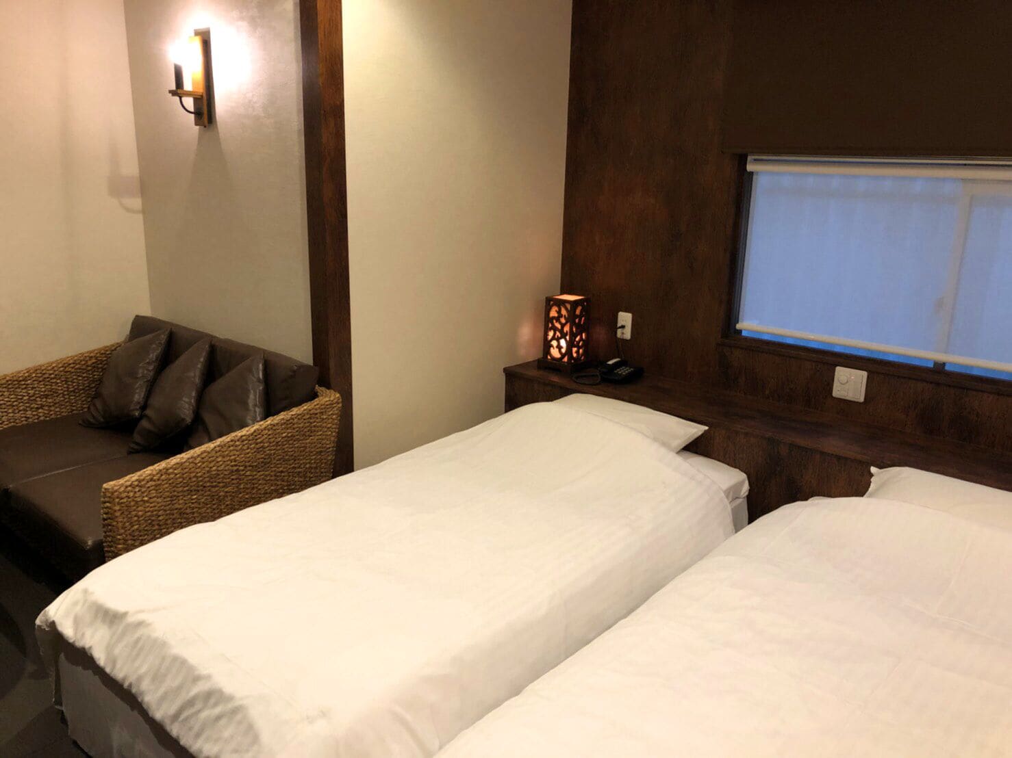 A hotel room with two single beds, a wicker sofa with cushions, a bedside table with a lamp, and a window with a view.