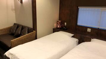 A hotel room with two single beds, a wicker sofa with cushions, a bedside table with a lamp, and a window with a view.