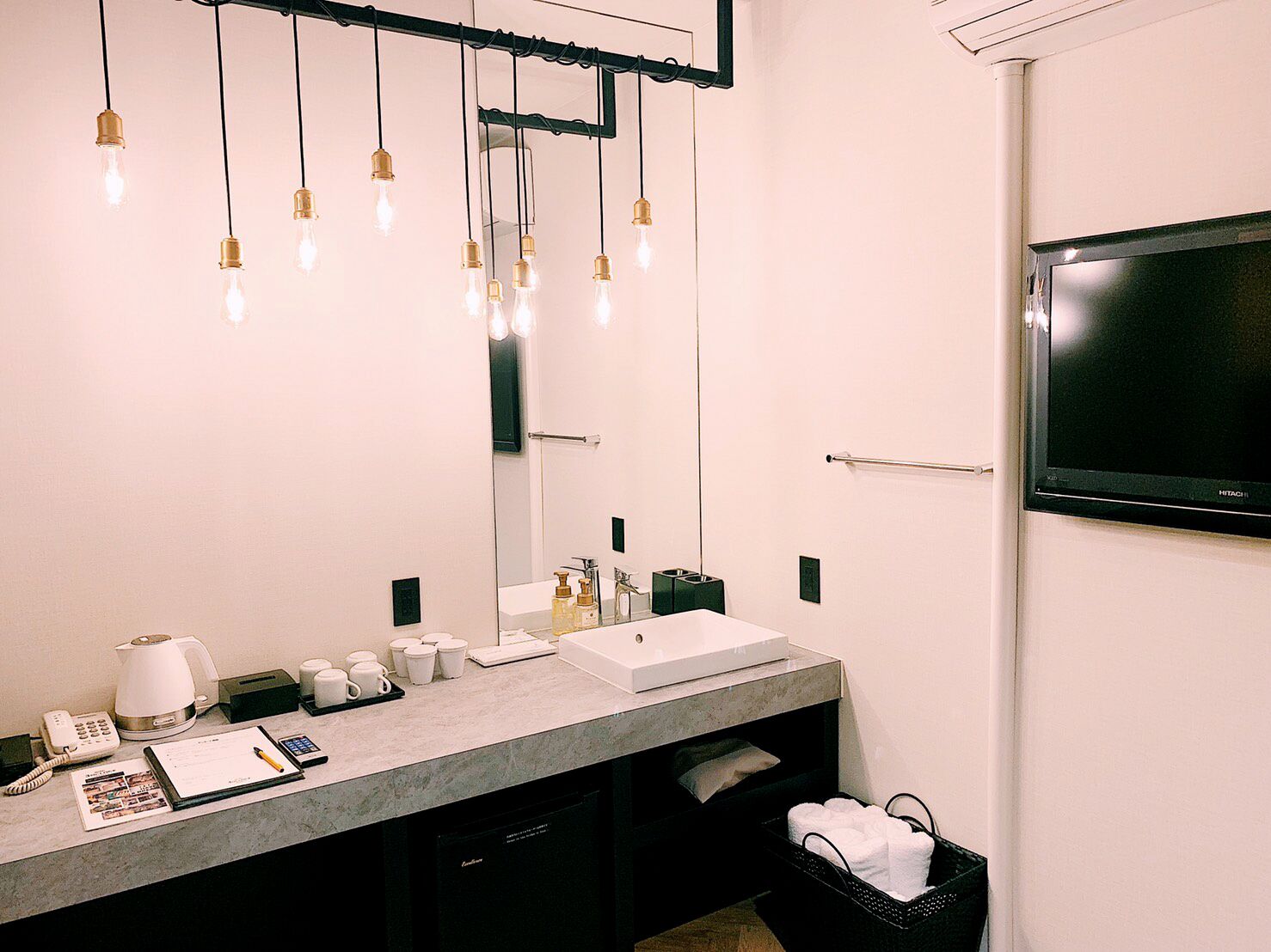 Un bany modern amb un mirall gran, un lavabo i un vàter. Hi ha llums penjants, una televisió i un cubell de brossa.