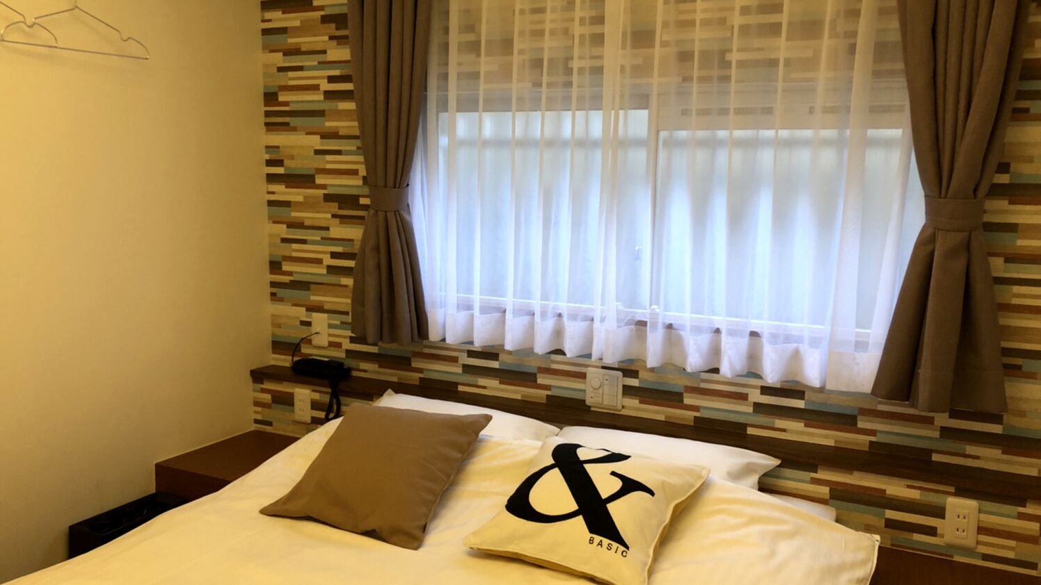 A hotel room with a bed, a window with curtains, a wall with a pixelated pattern, and a bedside table with a lamp.