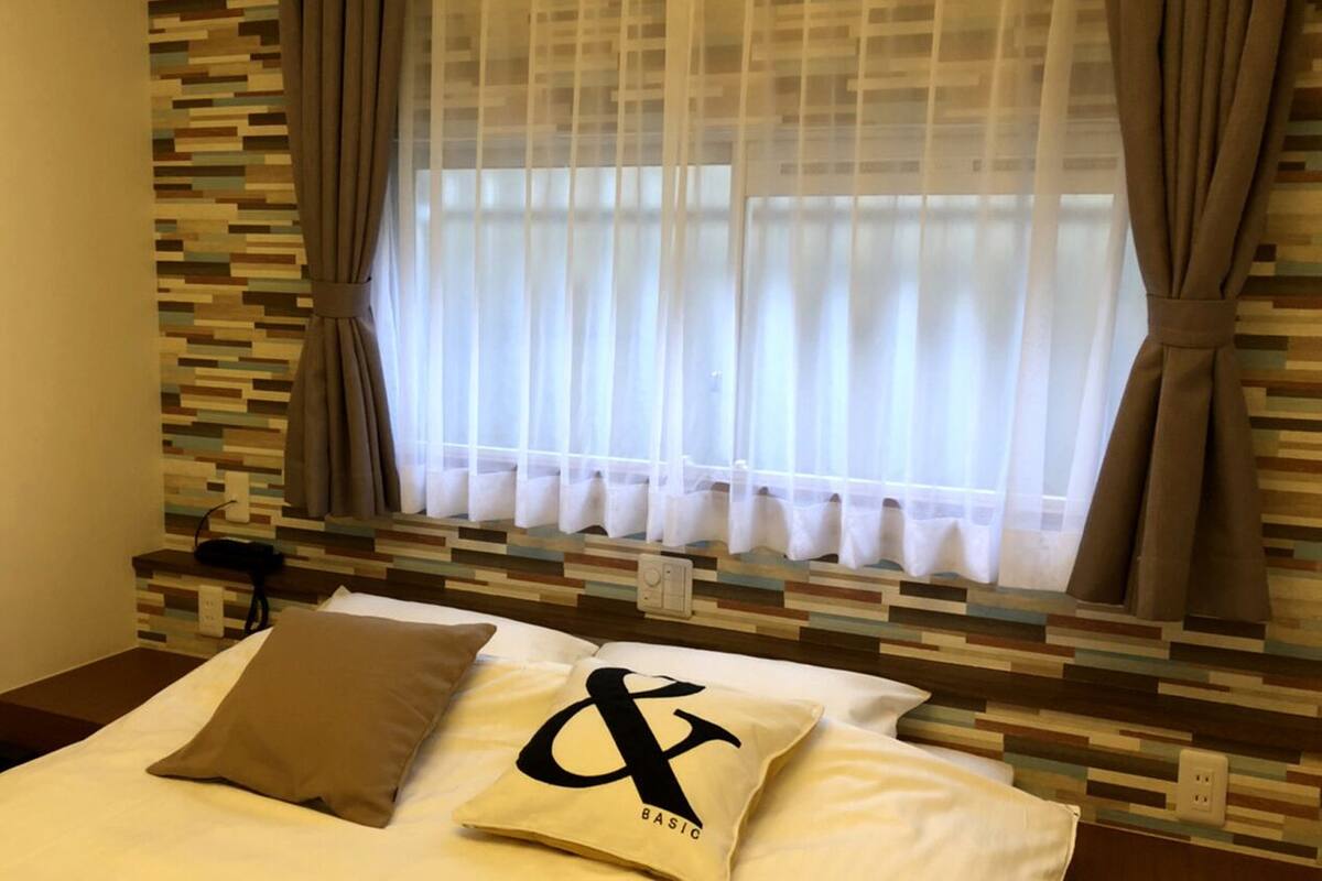 A hotel room with a bed, a window with curtains, a wall with a pixelated pattern, and a bedside table with a lamp.