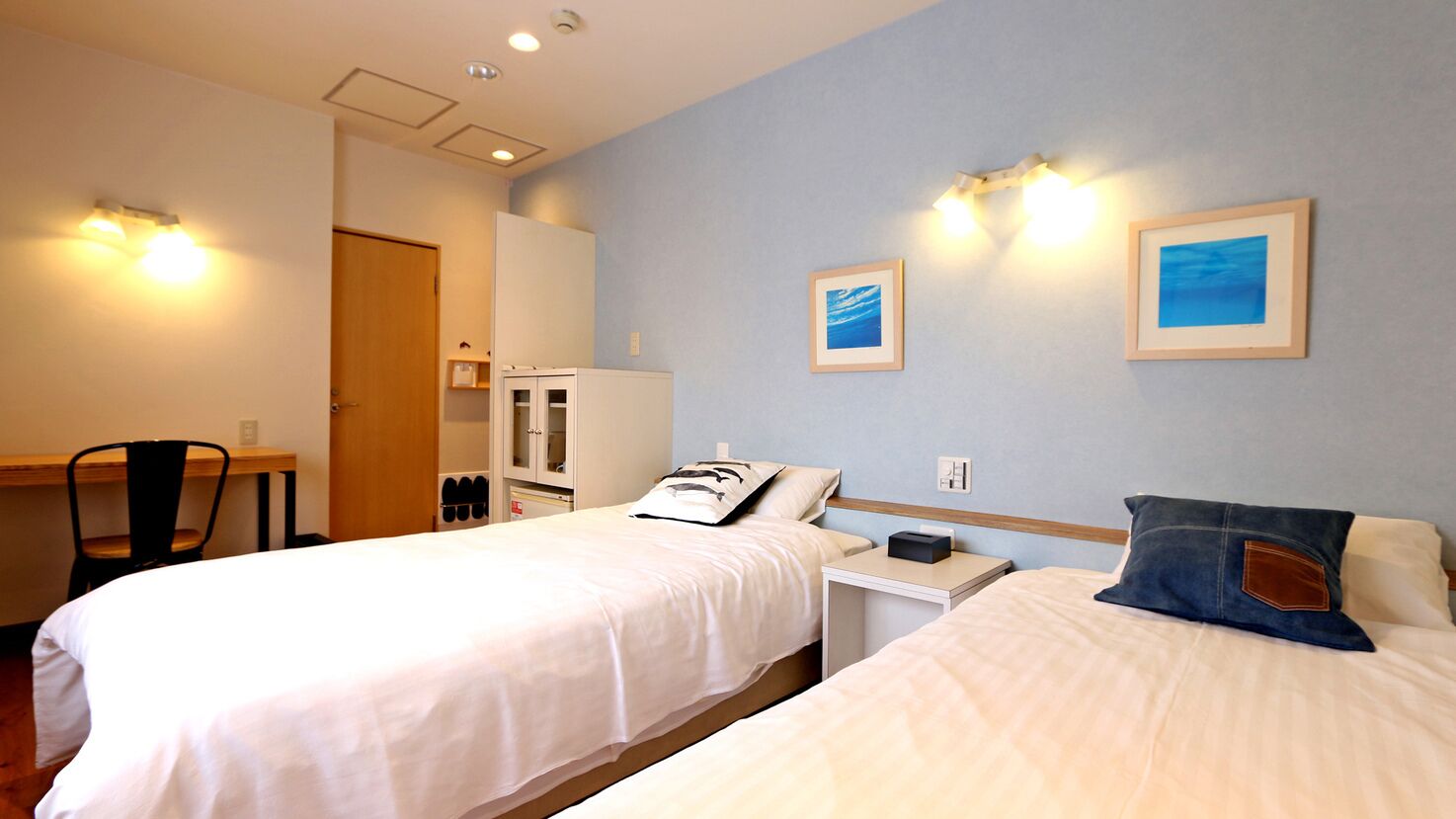 A hotel room with two beds, a desk, a chair, a wardrobe, and two framed pictures on the wall.