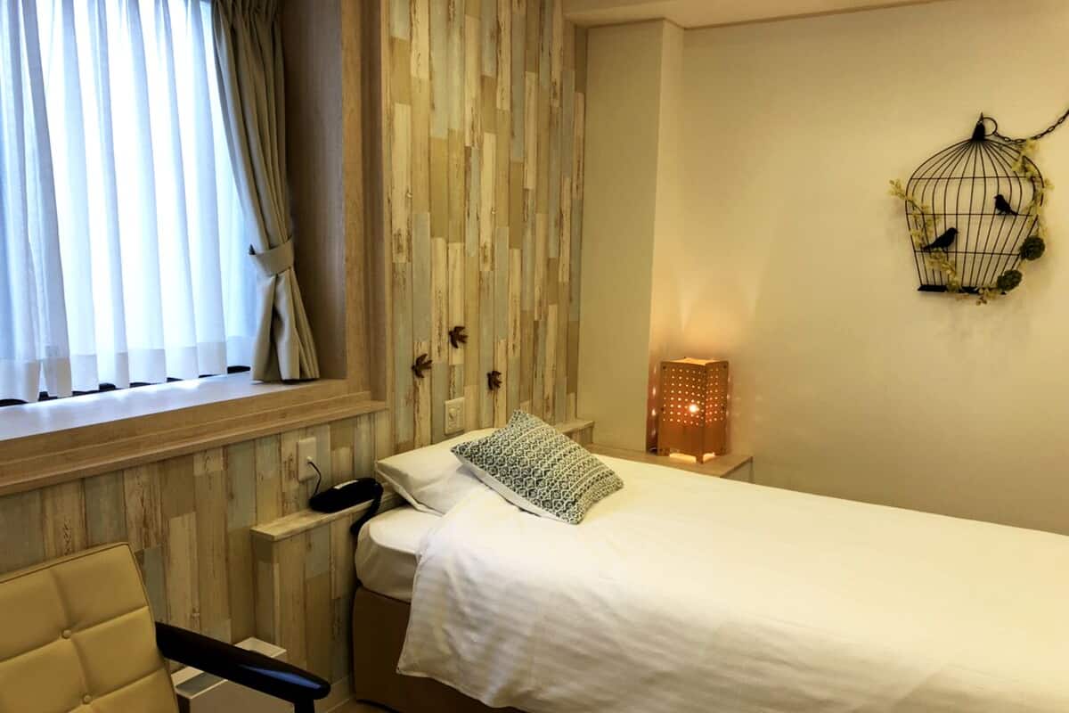 A bedroom with a bed, a chair, a bedside table, a wall-mounted light, and a birdcage decoration.