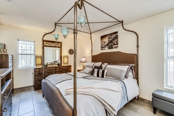 A bedroom with a four-poster bed, a dresser, a mirror, and a cityscape painting on the wall.
