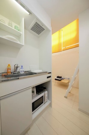 A compact kitchen with a microwave, sink, and storage cabinets. A small table and a chair are visible in the background.