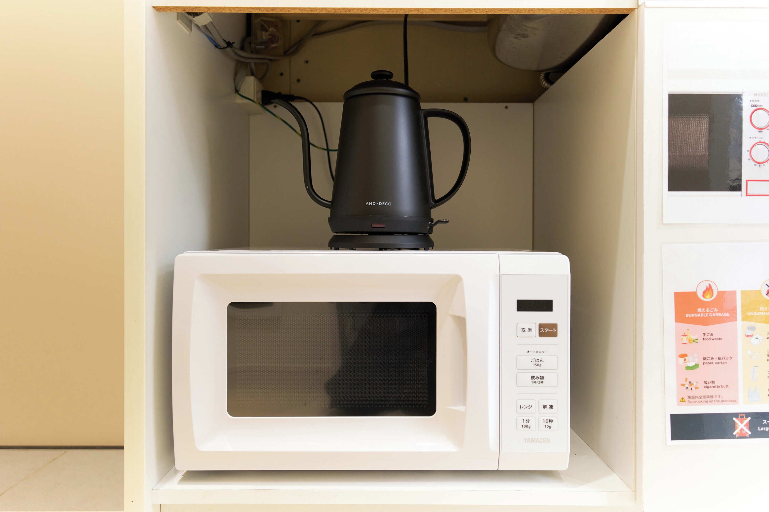 Een magnetron met een zwarte koffiepot erop, een bedieningspaneel met diverse knoppen, en een klein poster aan de rechterkant van de muur.