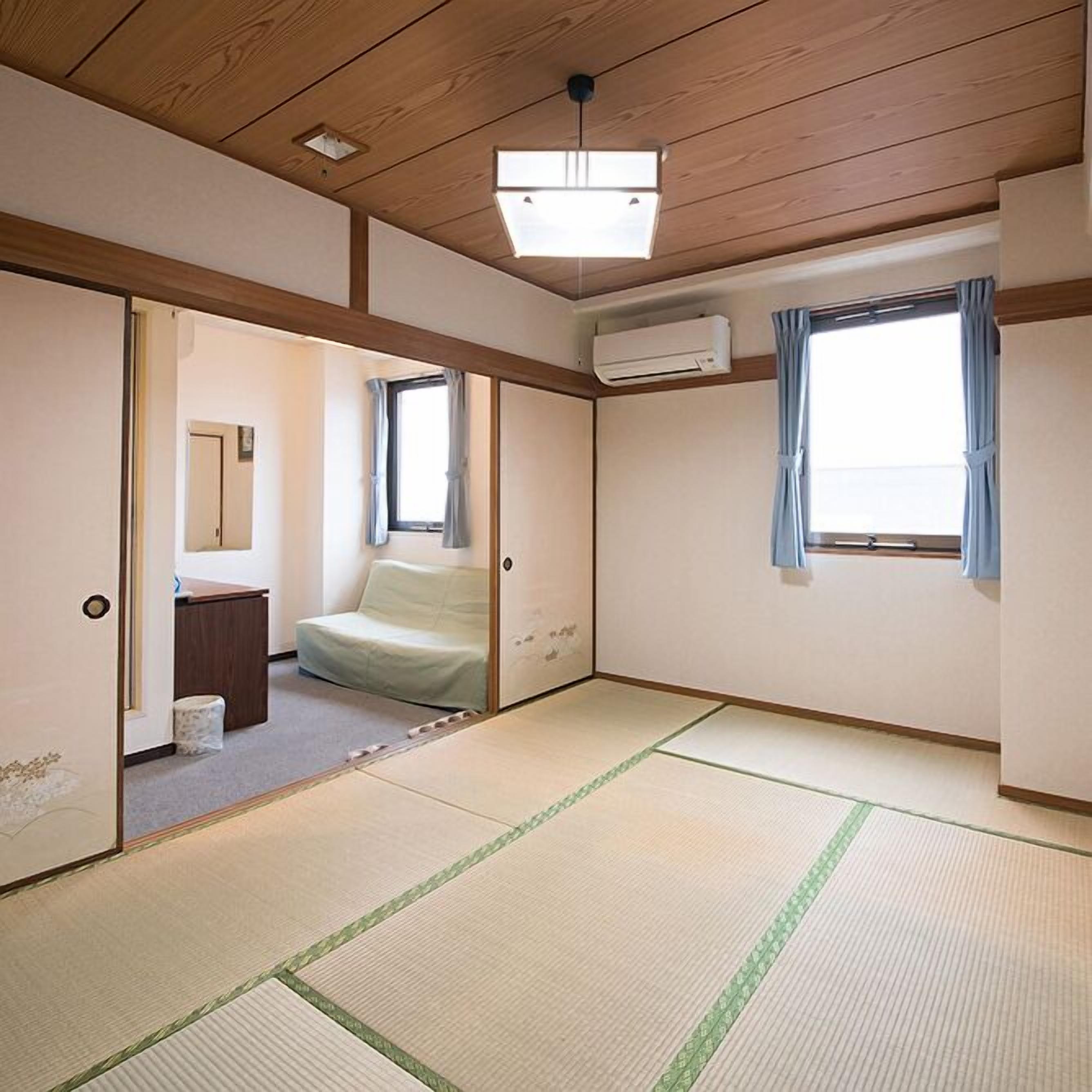 Une pièce de style japonais traditionnel, avec un sol recouvert de tatamis, une porte coulissante, un petit lit, une fenêtre avec des rideaux et un appareil de climatisation.