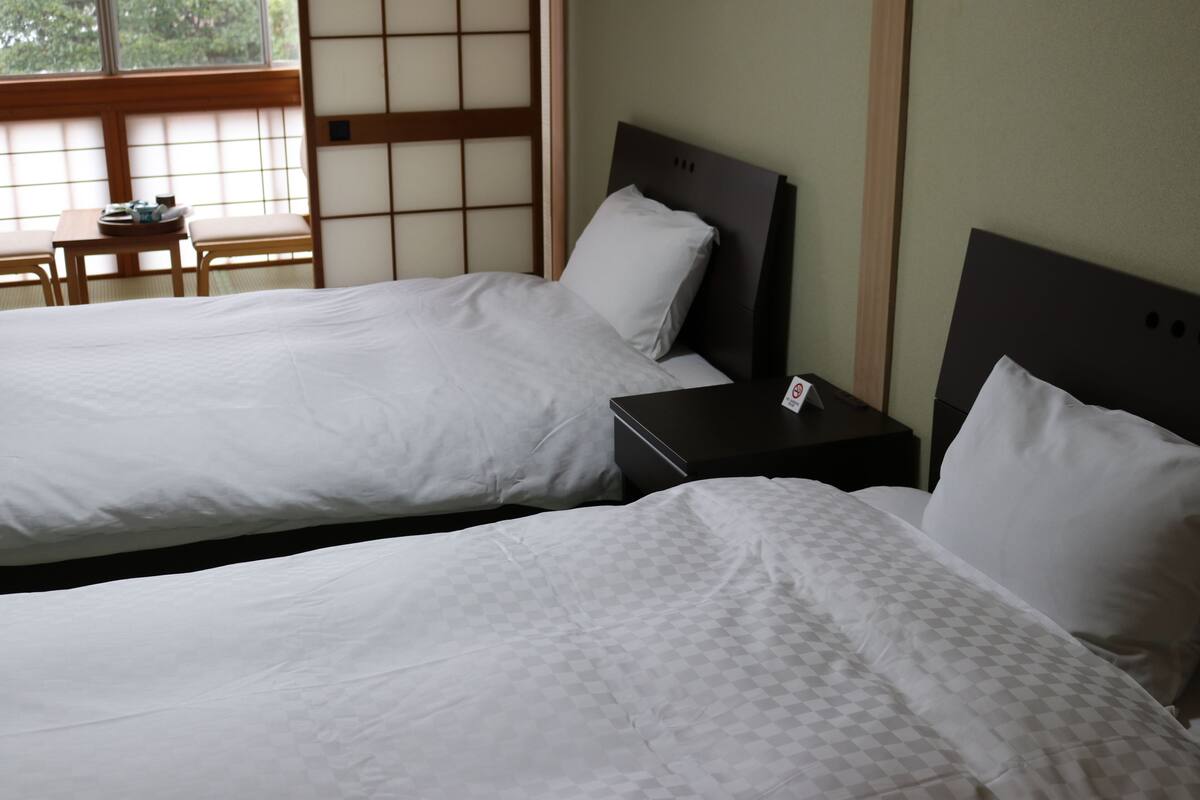 Deux lits simples avec du linge de lit blanc, une petite table en bois avec un plateau, et une vue sur une pièce traditionnelle japonaise avec un sol en tatami et des paravents shoji.
