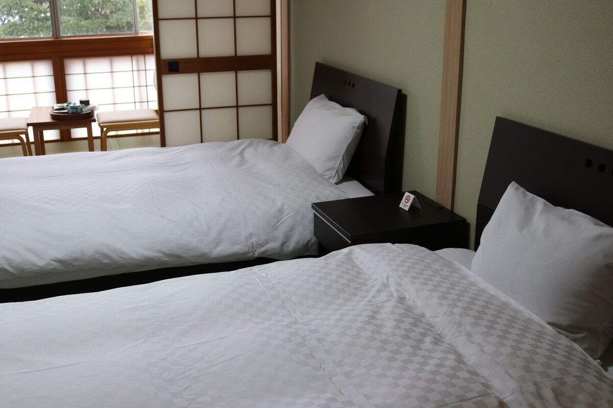 Deux lits simples avec du linge de lit blanc, une petite table en bois avec un plateau, et une vue sur une pièce traditionnelle japonaise avec un sol en tatami et des paravents shoji.