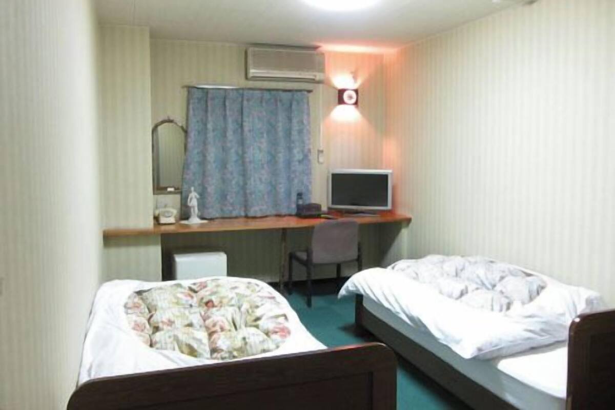 A room with two beds, a desk with a computer, a chair, and a wall-mounted lamp.