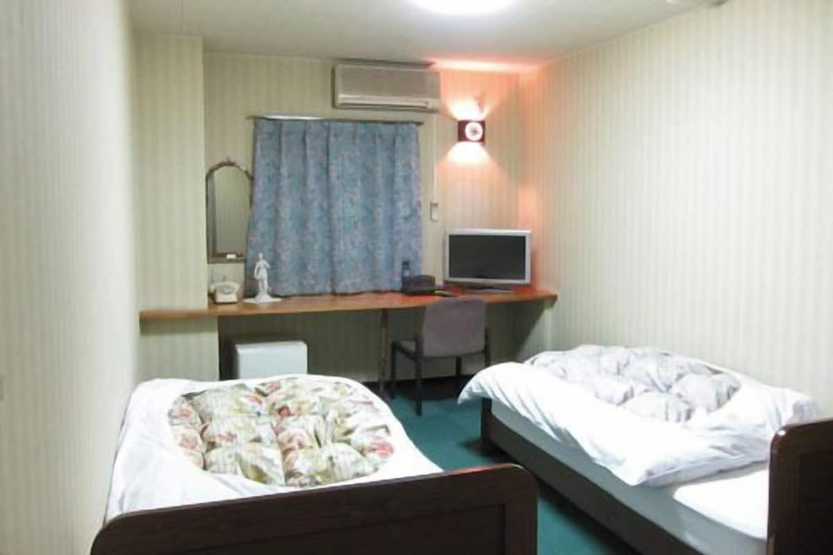 A room with two beds, a desk with a computer, a chair, and a wall-mounted lamp.