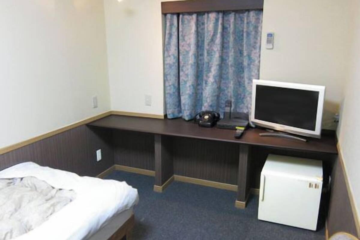 A hotel room with a bed, a desk with a computer monitor, a small refrigerator, and a window with curtains.