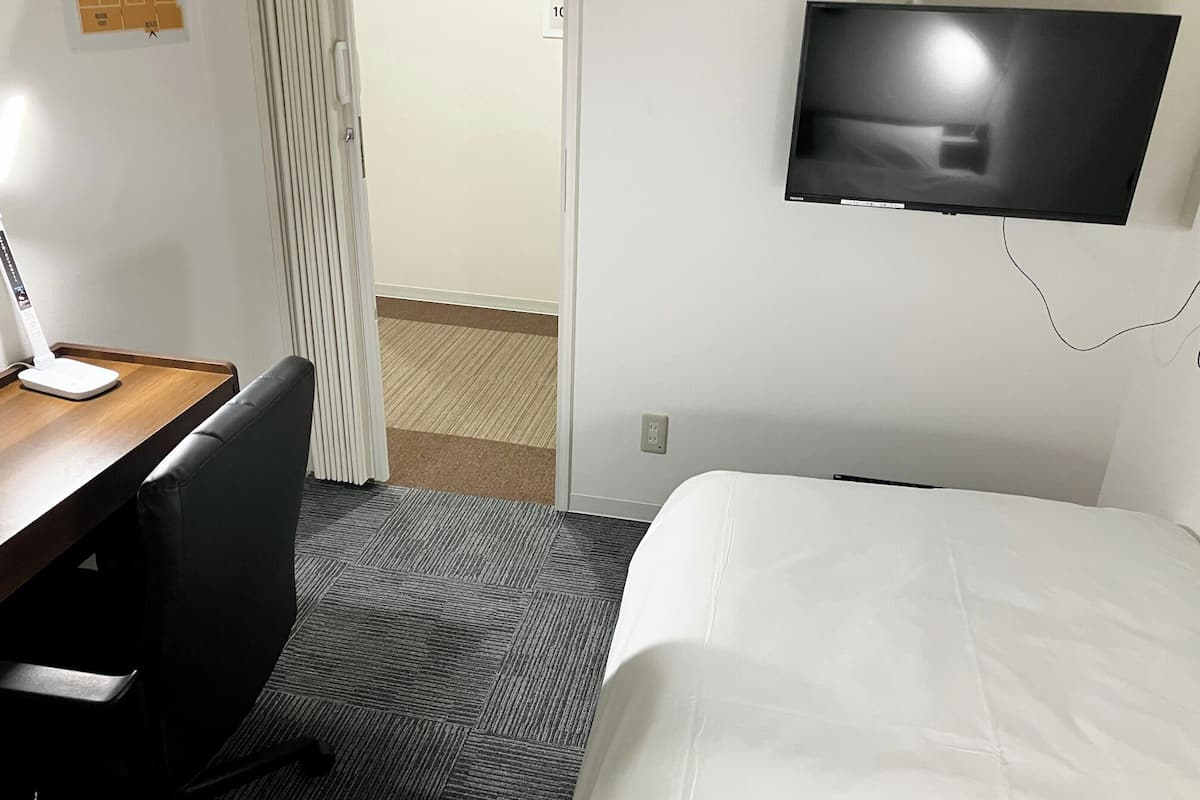 A hotel room with a bed, a desk with a lamp, a TV mounted on the wall, and a carpeted floor.