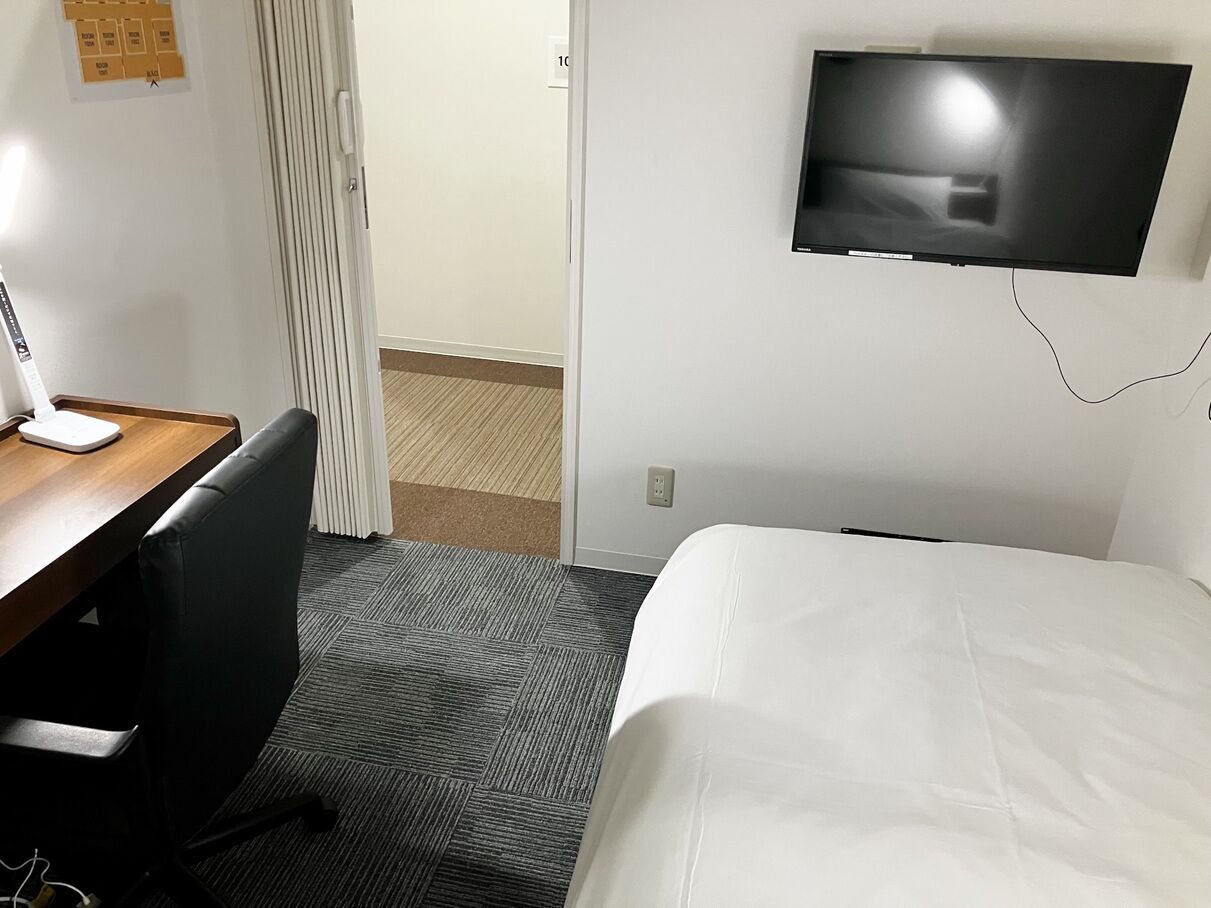 Hotellihuone, jossa on sänky, työpöytä tuolilla, seinälle asennettu televisio ja ovi, joka johtaa toiseen huoneeseen.