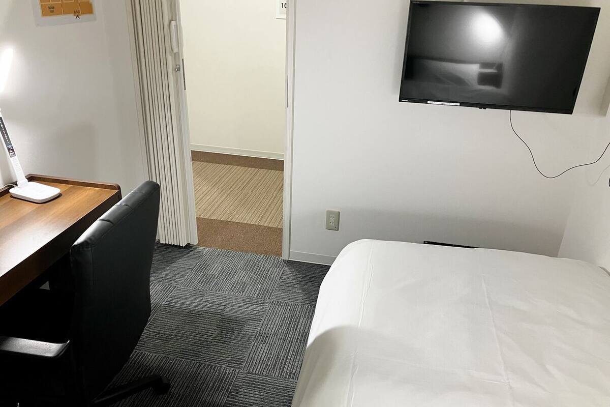 A hotel room with a bed, a desk with a chair, a television mounted on the wall, and a door leading to another room.