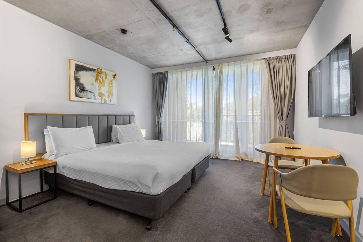 A hotel room with a bed, bedside table, lamp, TV, and a small table with chairs.