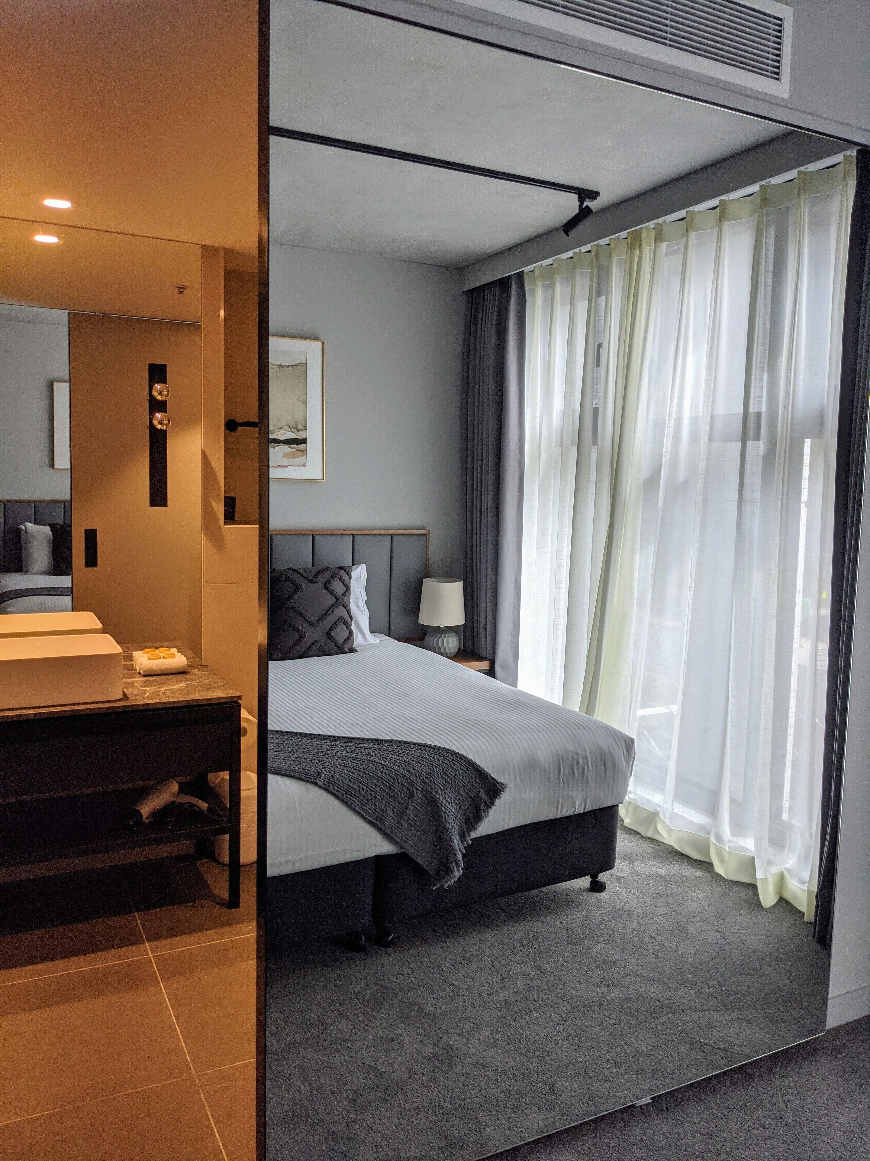 A modern hotel room with a large bed, a bedside table with a lamp, a bathroom with a sink and mirror, and sheer curtains on the window.