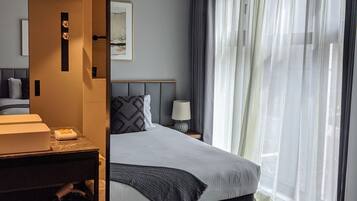 A modern hotel room with a large bed, a bedside table with a lamp, a bathroom with a sink and mirror, and sheer curtains on the window.