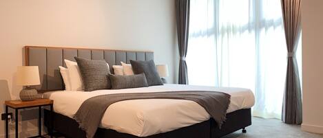 A modern bedroom with a large bed, a nightstand with a lamp, and a view of the outside through sheer curtains.