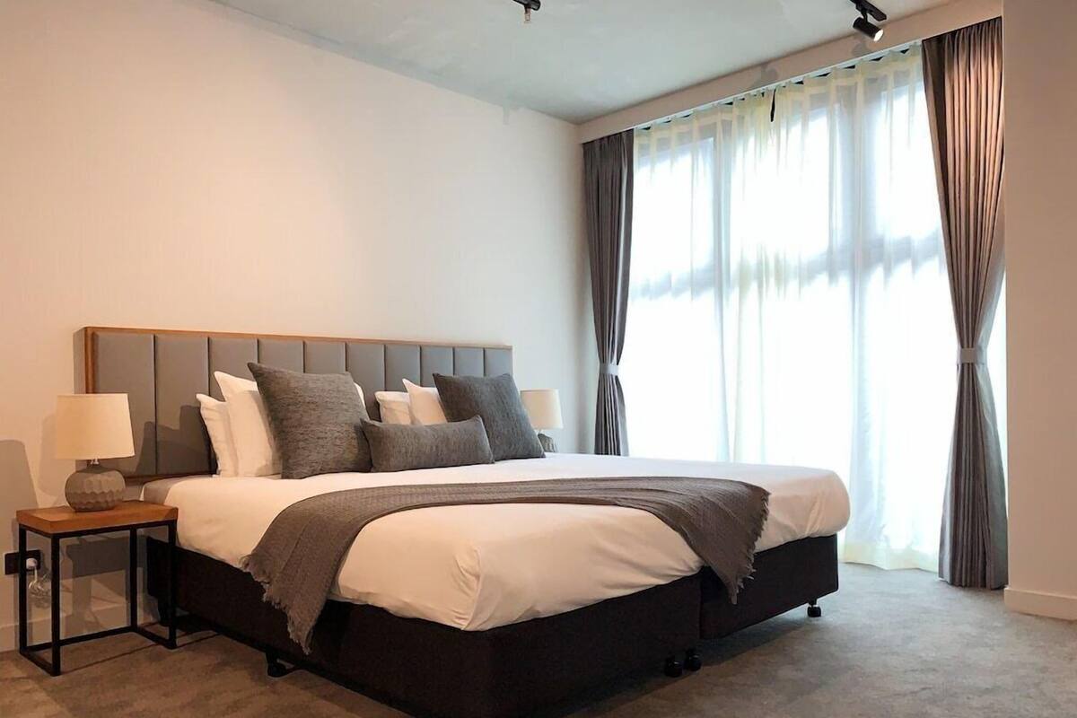 A modern bedroom with a large bed, a nightstand, a lamp, and a view of the outdoors through sheer curtains.