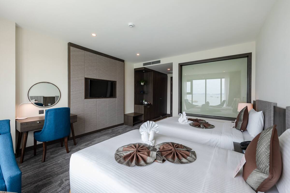 A hotel room with two beds, a desk with a chair, a TV, and a large window.