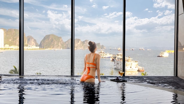 A woman in an orange swimsuit in a large indoor pool with a view of a body of water and boats. - Muong Thanh Grand Bai Chay Hotel (Bai Chay)