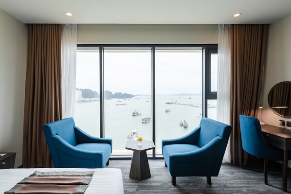 A hotel room with a large window, two blue armchairs, a small table with a vase, a bed, and a desk with a lamp.