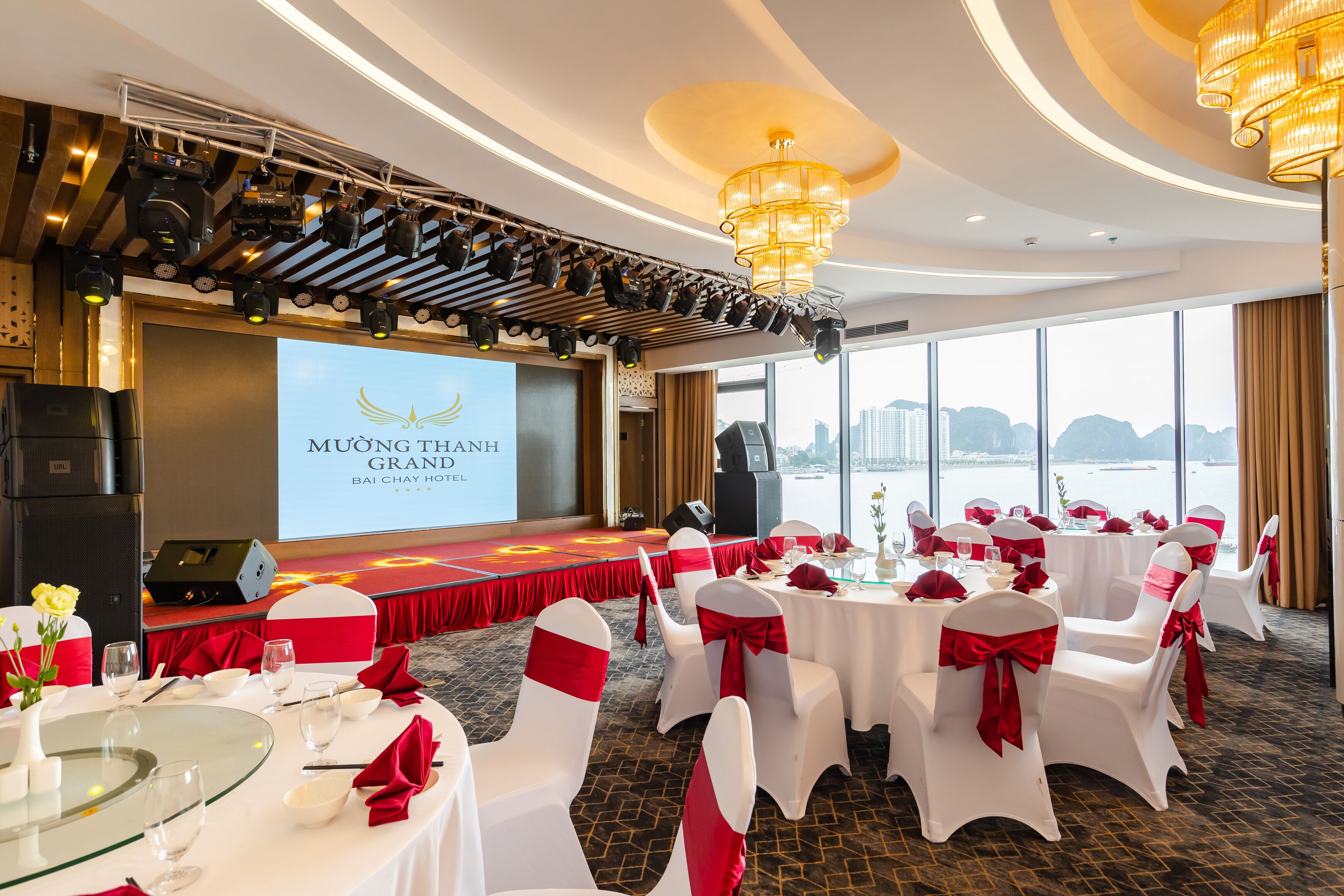 A banquet hall with round tables, white chairs with red sashes, a stage with a screen, and a city view through large windows.