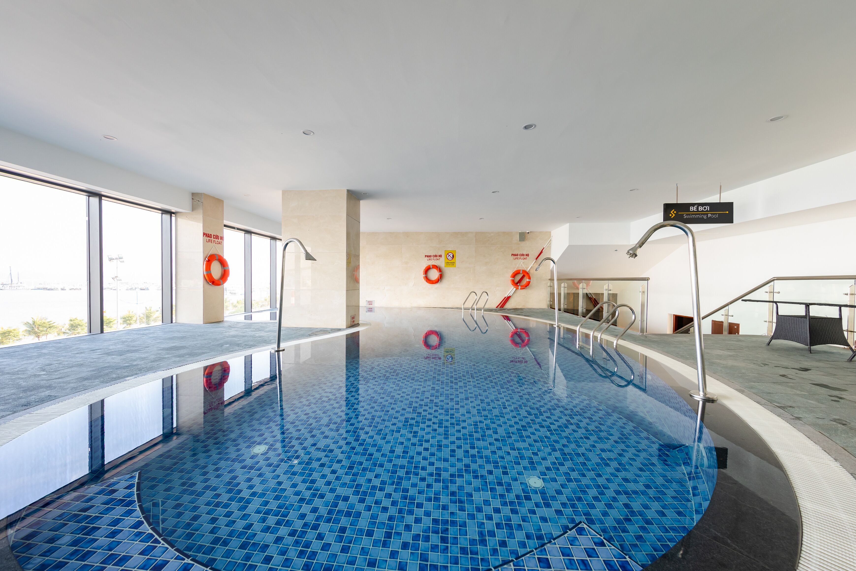 An indoor swimming pool with clear glass walls, a tiled floor, and a lifebuoy on the wall.