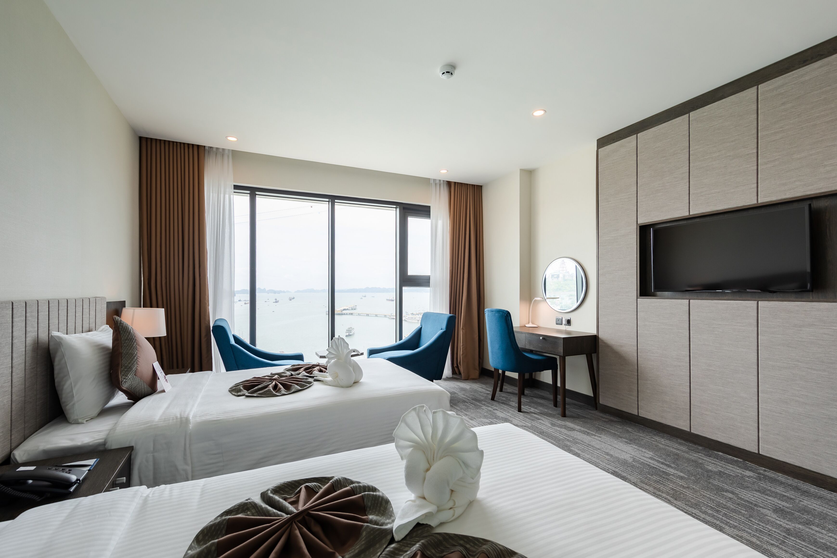A hotel room with two beds, a TV, a desk with a chair, and a view of the sea.