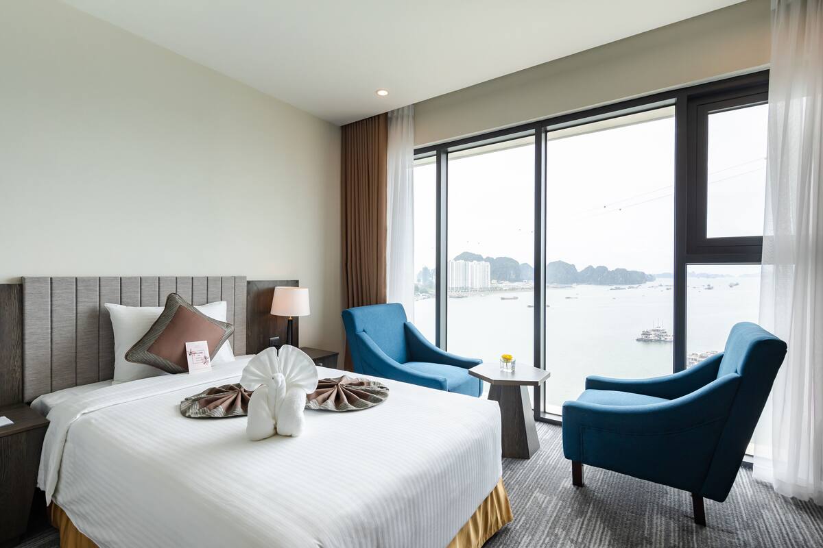 A hotel room with a large bed, two blue armchairs, a small table, and a view of a body of water through large windows.