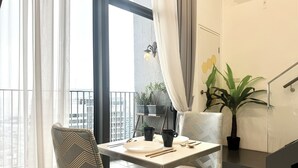 Ein Essbereich mit Tisch für zwei Personen, Stühlen, einem Blumentopf und Blick auf die Stadt durch große Fenster.