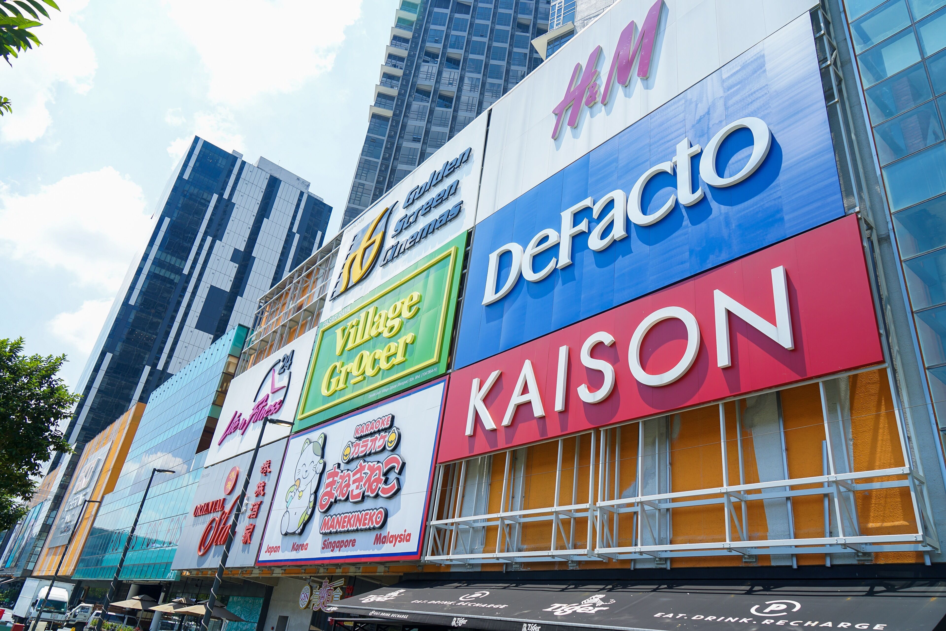 A building with multiple commercial signs, including 'Golden Green Themas', 'Village Grocer', 'Defacto', and 'Kaison'.