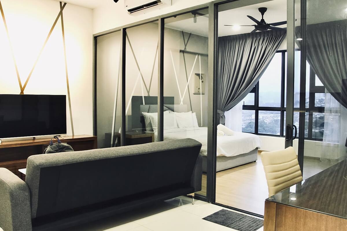 A modern hotel room with a sofa, a TV, a bed, a glass partition, and a view of the city.