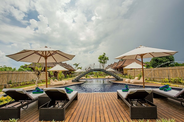 Terraza de madera con sillones de descanso y sombrillas con vista a una piscina y un puente.