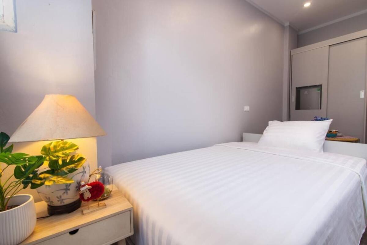 A neatly made bed with white bedding, a bedside lamp, a potted plant, and a small decorative item on a bedside table.