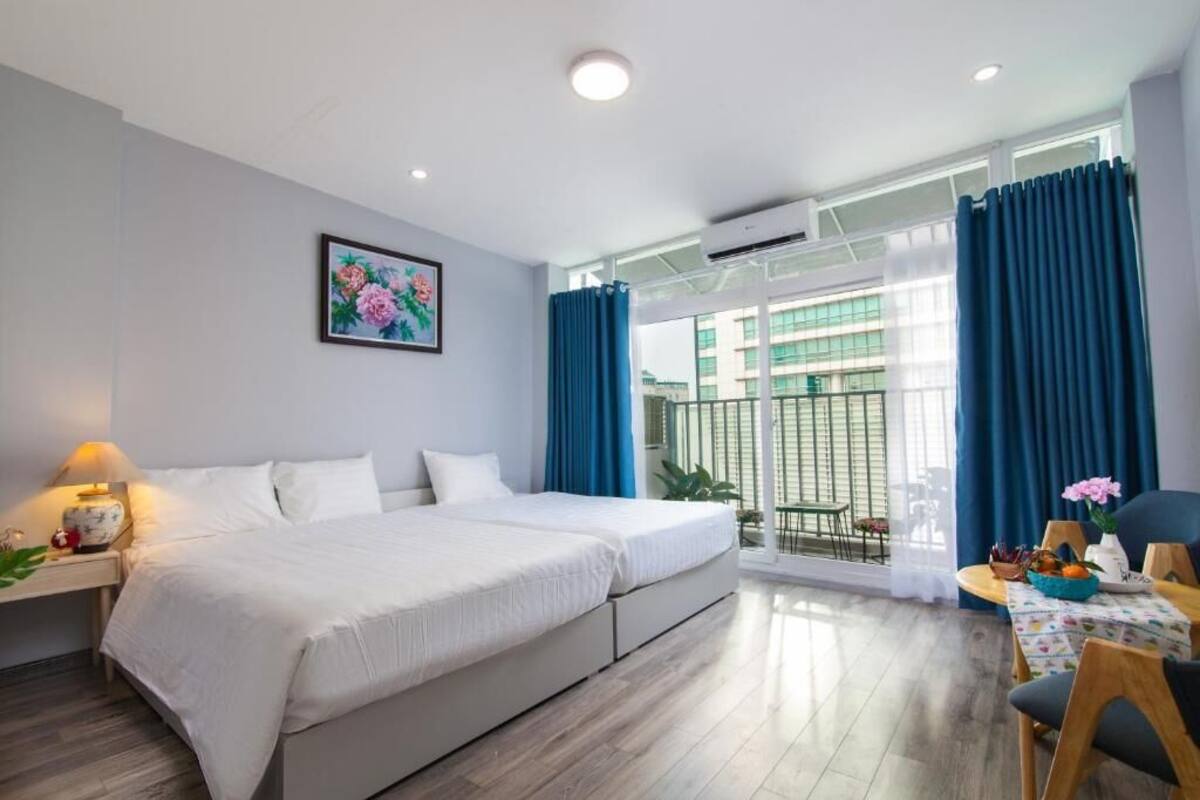 A modern hotel room with a large bed, a desk with a lamp, a dining area with a table and chairs, and a balcony with blue curtains.