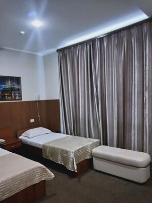 Quarto de hotel com duas camas de solteiro, cabeceira de madeira, uma janela grande com cortinas, luz noturna e vista para a cidade.