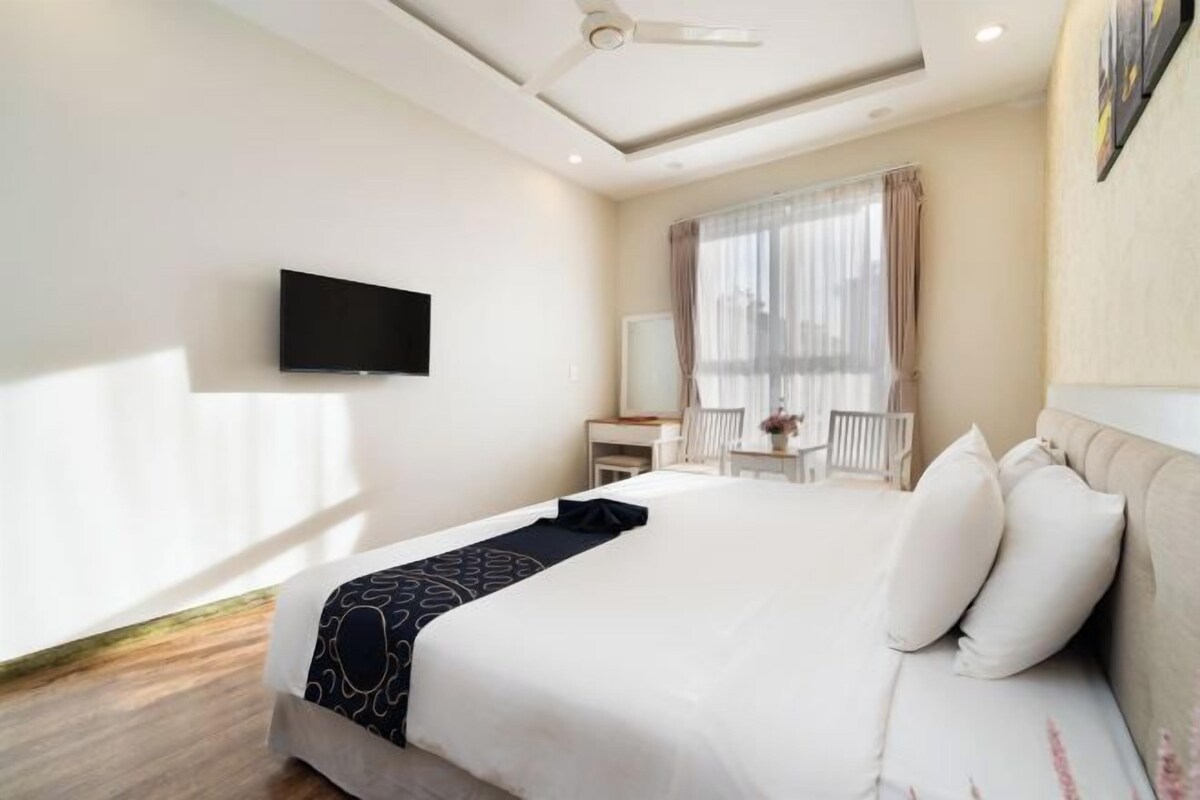 A hotel room with a bed, a flat-screen TV mounted on the wall, a desk with a chair, and a window with curtains.