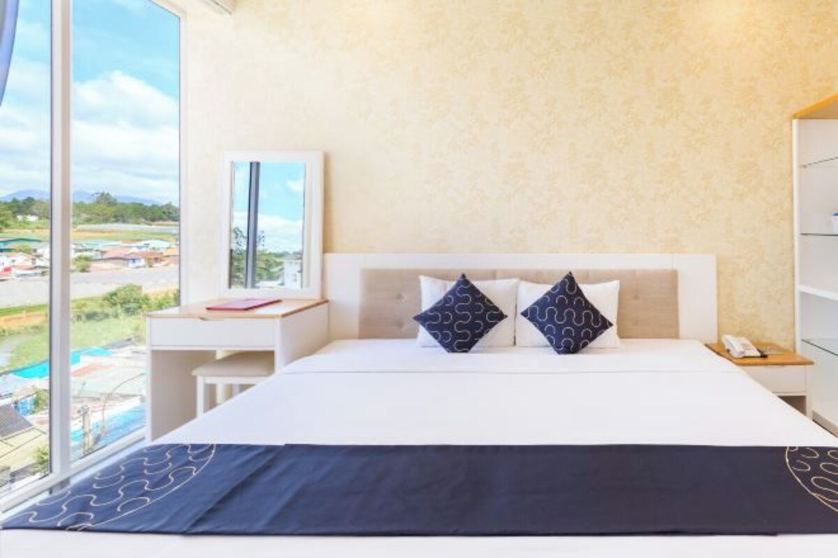 Habitación de hotel con una cama grande, una mesita de noche y vistas a una piscina y edificios.