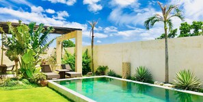 A rectangular pool with a tiled edge, surrounded by lush greenery and palm trees. There are lounge chairs and a pergola structure. - Private House YOSHINO (Miyakojima)