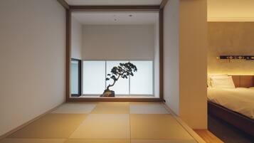 A minimalist hotel room with tatami flooring, a bed with white bedding, and a bonsai tree in the corner.