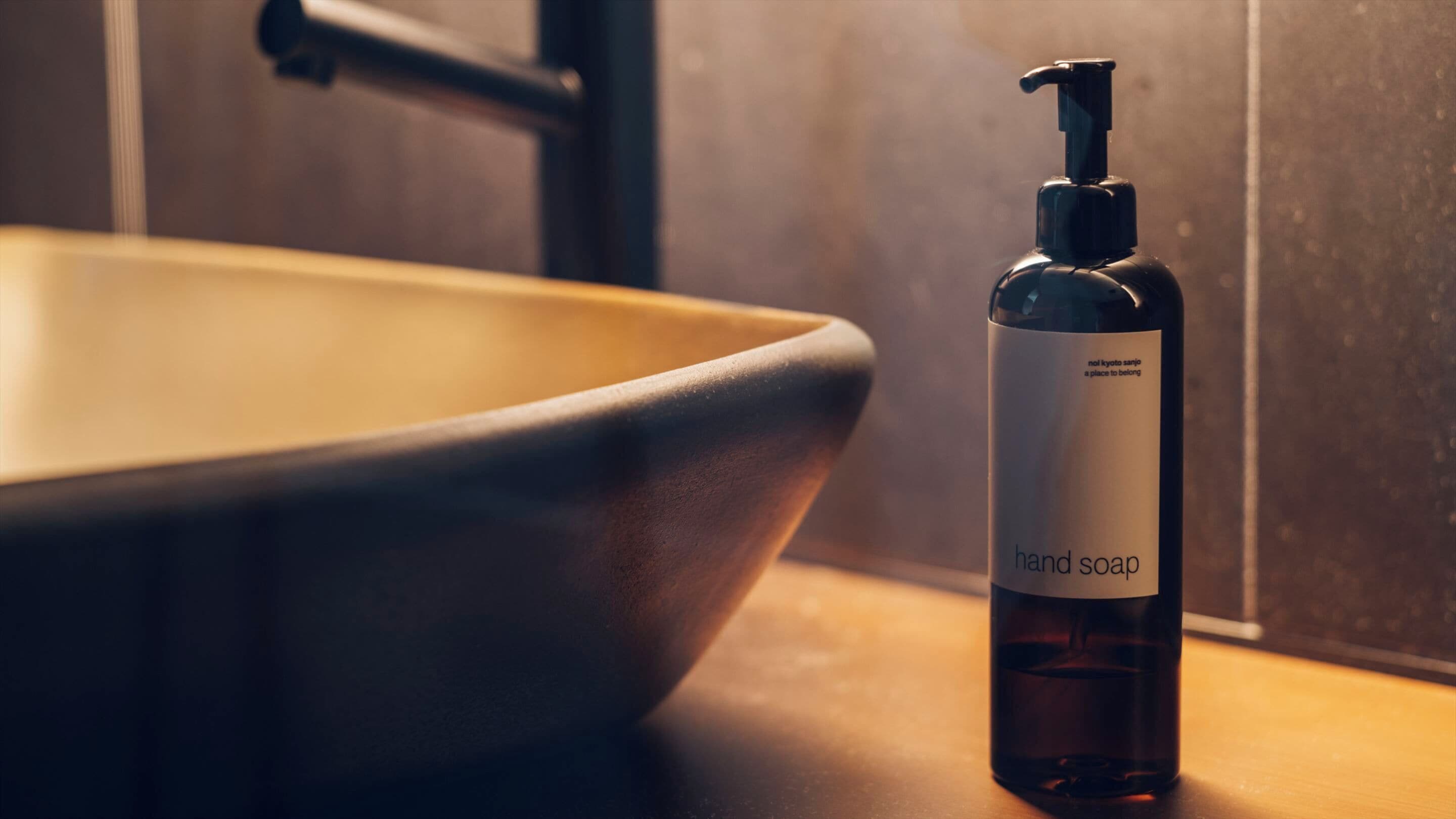 Un flacon de savon liquide pour les mains, doté d’un distributeur à pompe, est posé sur une surface en bois, à côté d’un grand évier en céramique de couleur beige.