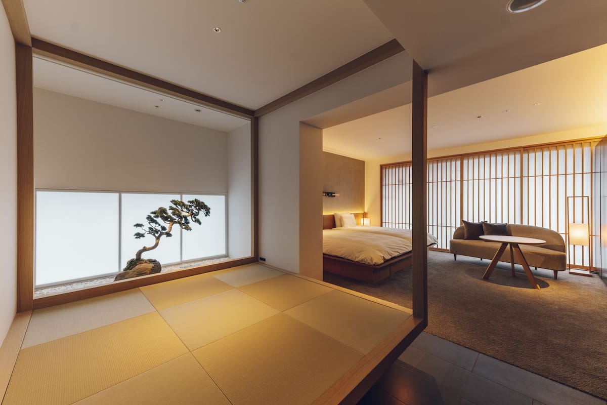 A modern hotel room with tatami flooring, a bed, a sofa, and a large window with a bonsai tree.