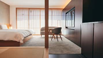 A modern hotel room with a bed, a chair, a TV, and a large window with blinds.