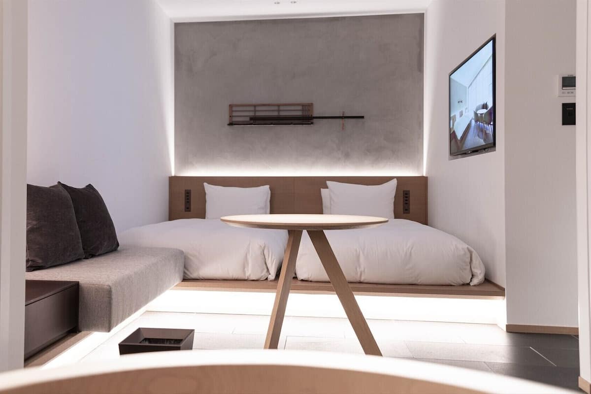 A modern hotel room with a bed, a sofa, a small table, and a TV mounted on the wall.