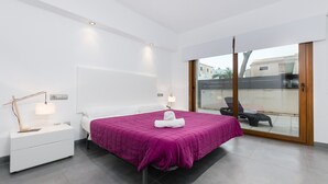 A modern bedroom with a large bed, a nightstand, a lamp, and a view of the outside through a sliding glass door. - YourHouse Cardinal (Can Picafort)